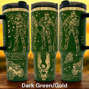 May include: Dark green travel mug with gold-colored illustrations of soldiers, vehicles, and text. The mug has a black handle and lid with a gold rim. The text includes "117", "UNSC", and a quote. The mug is designed with a military theme.