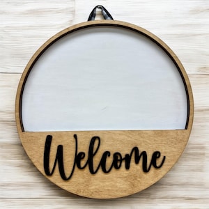 Peut inclure: Une pancarte ronde en bois avec un centre blanc et un bas brun. Le bas porte le mot "Bienvenue" écrit en lettres cursives noires.