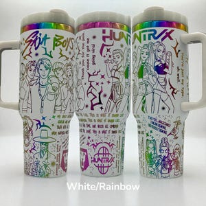 May include: Three white travel mugs with rainbow-colored illustrations and text. The mugs feature drawings of people and phrases like "Skit Boys" and "HuntraX." Each mug has a white handle and a white lid with a rainbow rim.