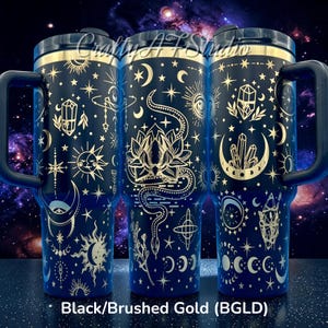 May include: Three black insulated tumblers with gold celestial designs, including moons, stars, and crystals. Each tumbler has a black handle and lid with a gold rim. The text "Crafty AT Studio" is visible on the top. The bottom of the image reads "Black/Brushed Gold (BGLD)".