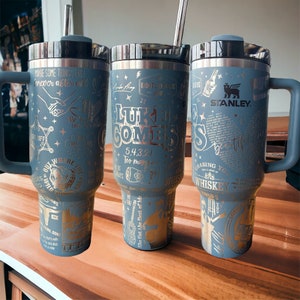 May include: Three stainless steel tumblers with lids and straws. The tumblers are blue with silver lettering and designs. The tumblers feature the text "Luke Combs" and "5.4.3.2.1 Too Many" on the middle tumbler. The tumblers are on a wooden surface.