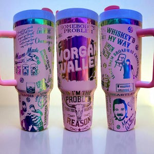 Może przedstawiać: Trzy różowe kubki termiczne z uchwytami, z różnymi grafikami i tekstem. Kubki mają jasnoniebieską pokrywkę i różowy uchwyt. Tekst zawiera zwroty takie jak "Morgan Wallen", "Whiskey'd My Way" i "I'm the Problem".