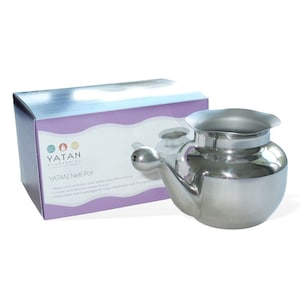 May include: A silver stainless steel neti pot with a rounded body and a spout. The pot is on a purple and white box with the text "YATAN AYURVEDICS" and "YATAN Neti Pot".