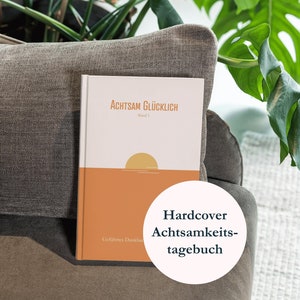 Könnte beinhalten: Ein Hardcover-Tagebuch mit dem Titel "Achtsam Glücklich" auf dem Cover. Das Cover ist weiß mit einer Sonnenuntergangsgrafik und der Text ist in Deutsch. Das Tagebuch liegt auf einem braunen Sofa mit einer grünen Pflanze im Hintergrund. Der Text "Hardcover Achtsamkeits-tagebuch" ist in einem weißen Kreis auf der rechten Seite des Bildes geschrieben.