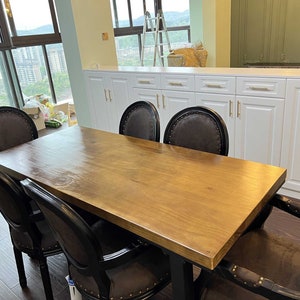 Puede incluir: Una mesa de comedor de madera con un acabado de madera natural y patas de metal negro. Seis sillas negras con tapicería de cuero marrón rodean la mesa. La mesa está ubicada en un comedor moderno con gabinetes blancos y una ventana grande.