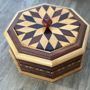 May include: An octagonal wooden box with a geometric design on the lid. The lid features a starburst pattern of dark and light wood, with a small, round, reddish-brown knob. The box's sides are a lighter wood with a darker wood trim.
