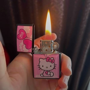 Può includere: Un accendino rosa Hello Kitty con una base nera e una fiamma che esce dalla parte superiore. L'accendino ha un design rosa e bianco con un palloncino a forma di cuore e le parole "Hello Kitty" sul lato.