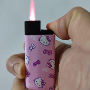 Può includere: Un accendino rosa con un motivo di Hello Kitty, con facce bianche di Hello Kitty e fiocchi rosa. L'accendino è acceso, mostrando una fiamma rossa. L'accendino è tenuto in una mano.