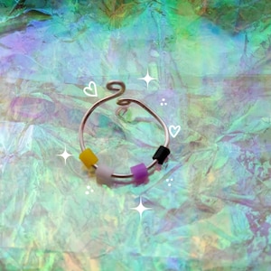 May include: A silver-colored nose ring with a spiral design at the top. The ring features a series of small beads in yellow, white, purple, and black. The background is a shimmering, iridescent material.