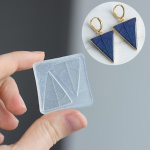 May include: Clear, square silicone mold with a leaf design, held in hand. The image also shows a pair of blue triangle earrings with gold-colored hoops. The earrings have a leaf texture and gold trim.