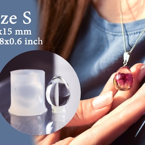 Może przedstawiać: Wisiorek z przezroczystej żywicy z różowym kwiatem w środku. Wisiorek jest zawieszony na srebrnym łańcuszku. Wisiorek ma 0,78 x 0,6 cala lub 20 x 15 mm. Wisiorek jest pokazany obok formy z przezroczystej żywicy.