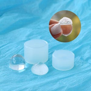 May include: Four clear silicone molds for resin casting. The molds are in different shapes, including a dome, a cylinder, and a half-sphere. The molds are on a blue surface.