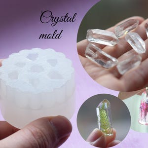 Può includere: Uno stampo in silicone bianco per realizzare oggetti in resina a forma di cristallo. Lo stampo ha otto cavità a forma di cristalli appuntiti. L'immagine mostra anche esempi di cristalli di resina finiti con fiori e vegetazione incorporati.