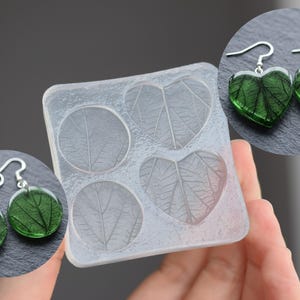 Puede incluir: Molde de silicona transparente con formas de hojas y dos pares de pendientes. Un par en forma de corazón y otro redondo, ambos con inclusiones de hojas verdes. Los pendientes tienen ganchos plateados.