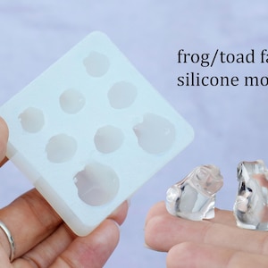 Könnte beinhalten: Eine transparente Silikonform mit acht Frosch- oder Krötenformen. Die Form wird in einer Hand gehalten. Zwei kleine, transparente Harzfrösche sind ebenfalls zu sehen.