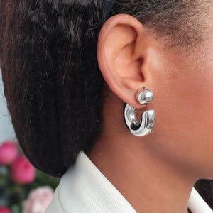 May include: A pair of shiny silver hoop earrings and a matching stud earring are displayed. The hoop earrings have a thick, rounded design, while the stud is a small, smooth sphere. The earrings are worn on a person's ear.