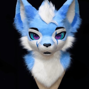 May include: A blue and white furry fox head with large ears, a white tuft of hair, and purple and teal eyes. The fox head has a black nose and is set against a black background. The fur is soft and fluffy.