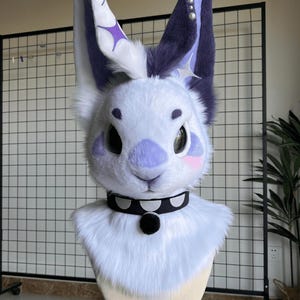 Puede incluir: Una cabeza de fursuit de conejo blanco y morado con orejas grandes y erguidas. Las orejas tienen detalles morados y en forma de estrella. La cara presenta ojos negros grandes, una nariz morada y mejillas rosadas. Un collar negro con un pompón negro alrededor del cuello.