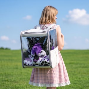 May include: A clear, square-shaped backpack with white trim containing a plush, furry creature with black and purple fur, pointed ears, and white paws. The backpack features a transparent front panel that displays the plush toy clearly inside.