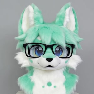 Peut inclure: Une tête de renard en peluche verte et blanche avec de grands yeux bleus portant des lunettes à monture noire. La fourrure est douce et moelleuse.