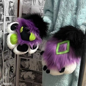 May include: A pair of purple and white furry paw hand puppets with black fur accents. One paw has a green diamond shape and the other has a green circle shape.