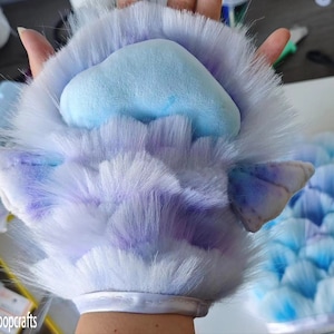 Op de afbeelding: Een close-up van een harige, lichtblauwe en paarse poot met een lichtblauw, wolkvormig kussentje. De poot heeft witte vachtaccenten en kleine, puntige oorachtige details. De tekst "Vibeloopcrafts" is linksonder zichtbaar.