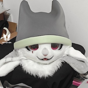 May include: A white furry animal head with a gray hat and a light green band. The hat has two pointy ears. The animal has red eyes and a pink tongue sticking out.