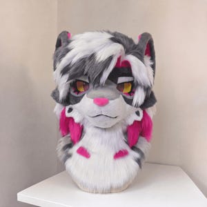 Puede incluir: Una cabeza de gato peluda con una combinación de colores blanco, gris y rosa. El gato tiene ojos amarillos, una nariz rosa y detalles rosas alrededor de las orejas y las mejillas. El pelaje está peinado con un patrón blanco y gris.