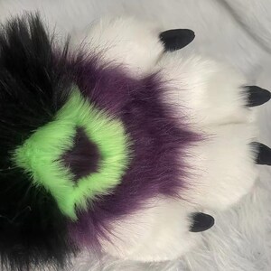 May include: A furry paw with black, purple, and green fur. The paw has black claws and is on a white fuzzy surface.