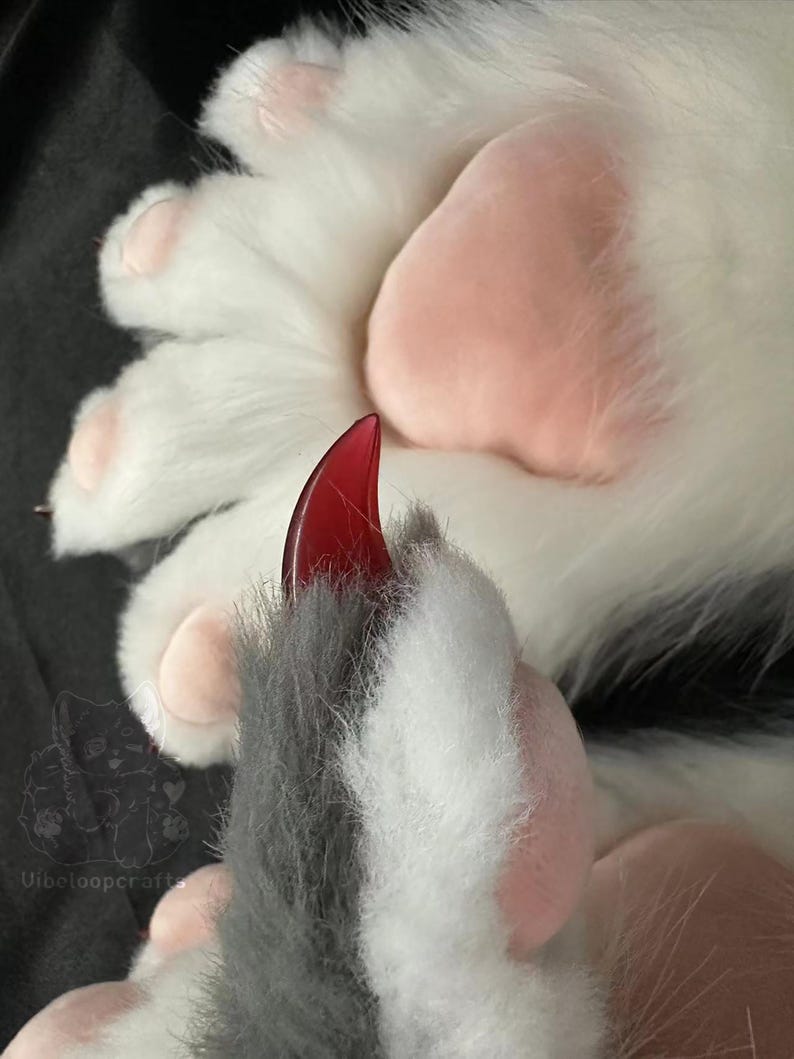 May include: Close-up of a plush paw with white fur and pink pads. A red claw extends from the paw. The paw is part of a larger costume or plush item. The image is well-lit and shows the texture of the fur.