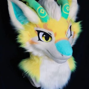 May include: A colorful furry head with a yellow, white, and teal color scheme. The head has large, expressive eyes, teal-colored horns, and a fluffy white and yellow mane. The nose is teal, and the ears are white with teal interiors.