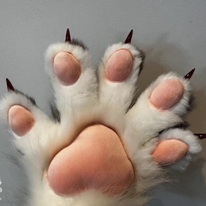 May include: A large, plush paw in white fur with grey detailing and pink pads. It has sharp, burgundy-coloured claws. The design resembles a large animal paw, possibly for a costume or novelty item. The paw is likely a prop.