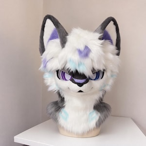 Può includere: Una maschera a testa di animale in peluche con lunga pelliccia sintetica morbida bianca e grigia. Il design include grandi orecchie a punta con accenti viola, occhi viola brillanti e segni azzurri sulla fronte, sulle guance e sul petto con naso nero.
