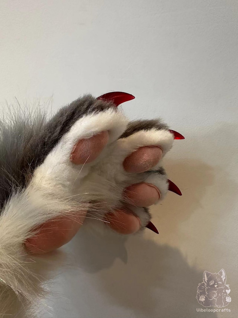 May include: A close-up shot of a furry paw featuring pink pads and striking red claws. The fur is a blend of grey and white, with a soft, plush appearance. The image is well-lit, accentuating the details of the paw and claws. The theme suggests fantasy or cosplay.