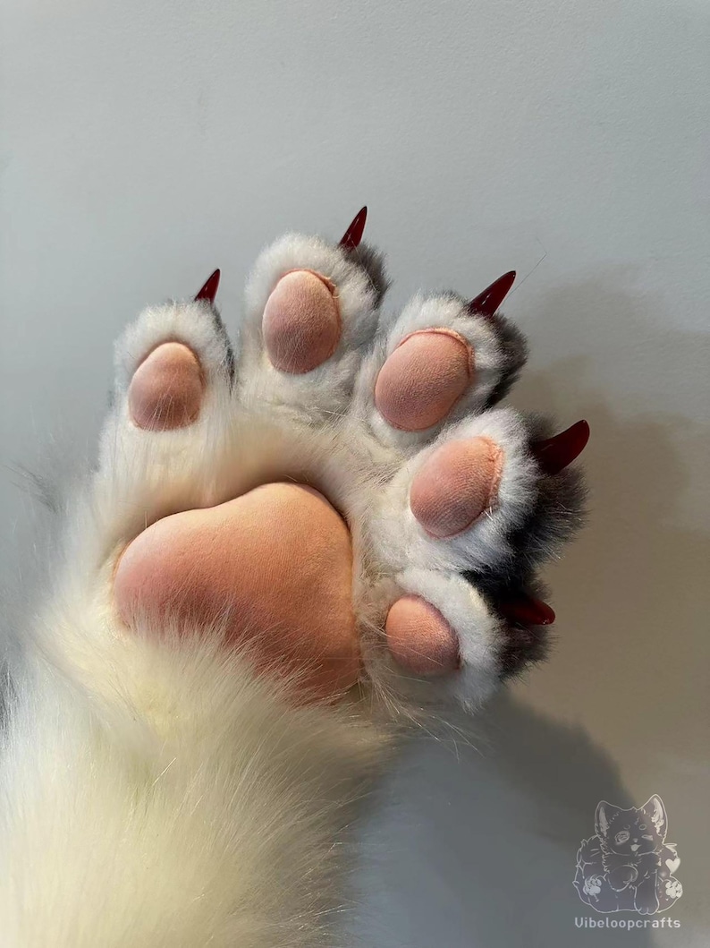 May include: A close-up of a plush paw featuring white fur, grey detailing, and pink pads. The paw has five pink pads and sharp, red claws. The image depicts a furry, animal-like paw.