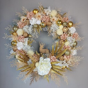 Op de afbeelding: Een gouden en crèmekleurige kerstkrans met een houten rendieruitsparing in het midden. De krans is versierd met kunstbloemen, bessen en dennenappels.