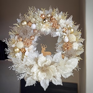 Op de afbeelding: Een witte en gouden kerstkrans met een hert silhouet in het midden. De krans is versierd met witte en gouden bloemen, bessen en ornamenten.