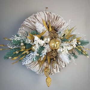 Op de afbeelding: Een beige fluwelen kerstkrans versierd met gouden en witte ornamenten, dennentakken en dennenappels.