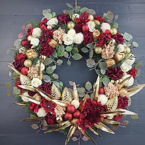 Op de afbeelding: Een kerstkrans met rode en gouden accenten. De krans is gemaakt van groen, rode en witte bloemen, gouden ornamenten en gouden dennenappels.