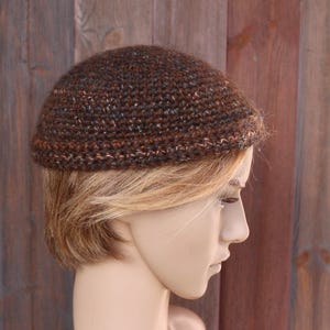Puede incluir: Un gorro marrón tejido a ganchillo con motas de color naranja, azul y blanco. El gorro tiene forma de cúpula y parece hecho a mano. El gorro se muestra en una cabeza de maniquí con cabello rubio. El fondo es una pared de madera.