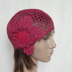 Puede incluir: Un gorro de ganchillo de color burdeos oscuro con un gran diseño floral y cuentas rojas. El gorro está hecho con un punto de calado delicado.
