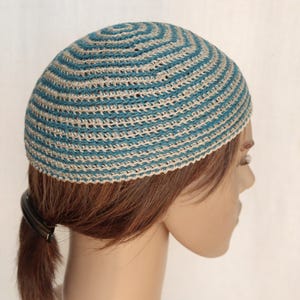 Puede incluir: Un gorro de calavera de ganchillo a rayas turquesa y blanquecinas. El sombrero está hecho de un hilo ligero, con un diseño circular. El gorro se muestra en la cabeza de un maniquí, con la parte posterior de la cabeza y el pelo visibles.