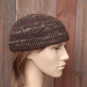 May include: A brown, hand-knitted beanie hat with a textured appearance. The hat features a mix of brown, tan, and white yarns, creating a speckled effect. The brim has a thin white stripe. The hat is displayed on a mannequin.