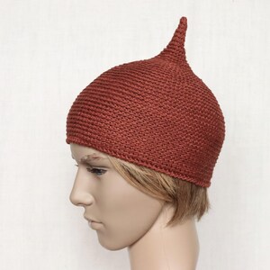 May include: A rust-colored, knitted hat with a pointed top. The hat is made of a textured yarn and has a close-fitting design. The hat is shown on a mannequin head.