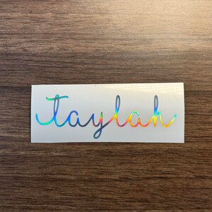 Könnte beinhalten: Ein weißer Aufkleber mit dem Namen "Taylah" in einer regenbogenfarbenen holografischen Schrift.