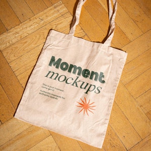 May include: A beige tote bag with the text "Moment mockups" in green lettering. The bag also features a small orange sun graphic and the text "This is your moment, come get it! Authentic moments for authentic brands."