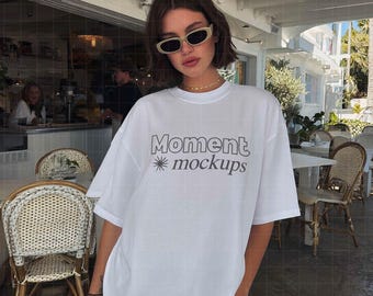 Op de afbeelding: Een wit t-shirt met de grijze tekst "Moment mockups", gedragen met een spijkerbroekshort. De persoon draagt een zonnebril en een gouden ketting. De achtergrond toont een restaurant met zitplaatsen buiten.