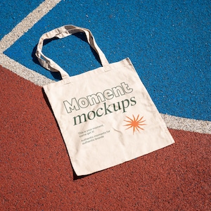 May include: A beige tote bag with the text "Moment mockups" and the tagline "This is your moment, come get it! Authentic moments for authentic brands". The bag is lying on a red and blue surface.