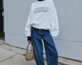 May include: A person wearing a white sweatshirt with the text "Moment mockups", dark blue jeans, and white sneakers. The person is also wearing a white baseball cap with a red brim, black sunglasses, and a small tan handbag.