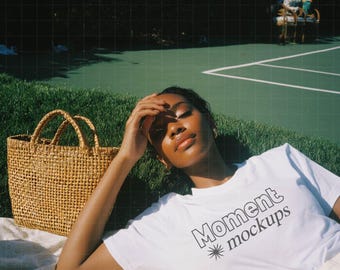 May include: A white t-shirt with the text "Moment mockups" in black, worn by a person lying on a blanket. A woven basket and a yellow glass are nearby. The scene is set outdoors on a sunny day.